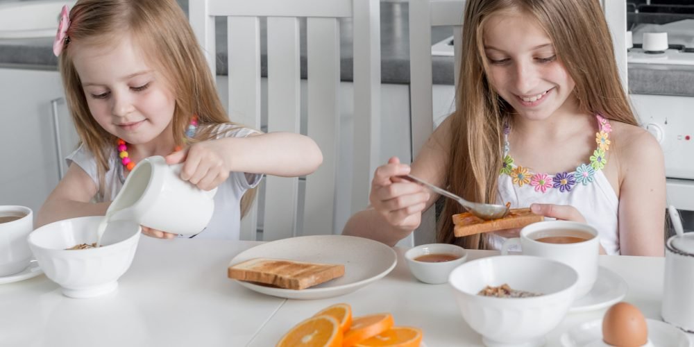 Two lovely sisters at the table eating healthy breakfast with toasts with honey and milk